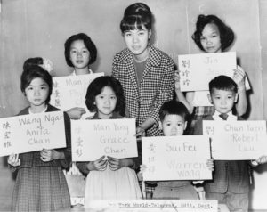 April Lou, teacher at Public School 1 in lower Manhattan, with six children who had recently arrived from Hong Kong and Taiwan in 1964, holding up placards giving his or her Chinese name (both in ideographs and in transliteration) and the name to be entered upon the official school records. Photo credit: Fred Palumbo.